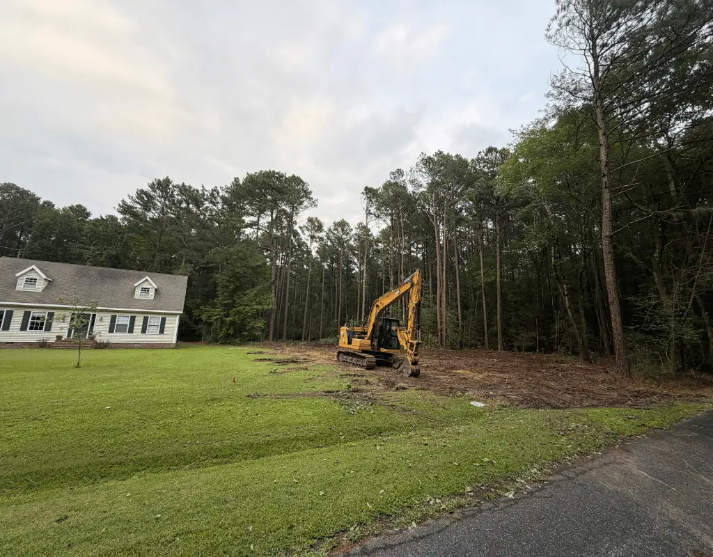 Excavator clearing brush along a pine tree line beside a house on the Delmarva Peninsula